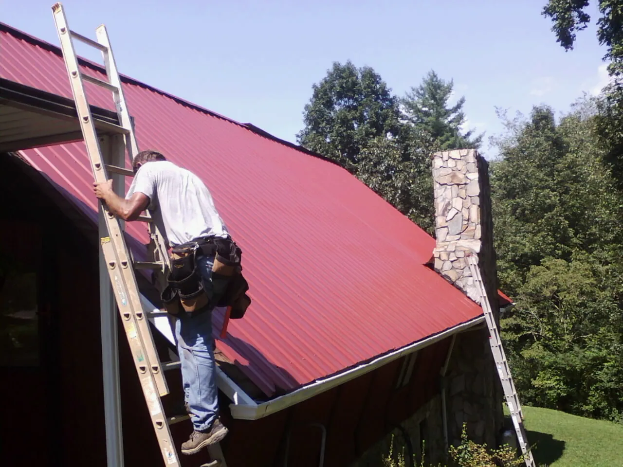 Roofer on a ladder beside a red metal roof and stone chimney — Soffit & Fascia Repair by Roofing Brothers in Vermontville